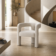 A modern upholstered accent chair in a textured light beige fabric, positioned in a minimalist, warm-toned interior with a sweeping concrete staircase in the background.