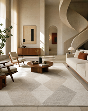 A large modern area rug with a light beige and off-white geometric diamond pattern, styled in a contemporary, sunlit living room with a wooden coffee table, armchair, and a spiral staircase in the background.