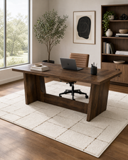 A dark brown wooden writing desk in a modern home office setting, styled with a laptop, books, a black chair, a potted olive tree, and a large cream-colored rug on a wood floor.