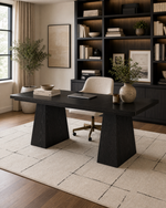 A modern dark wood writing desk with blocky pedestal legs, styled in a home office setting with a cream swivel chair, rug, and a large built-in bookshelf in the background.