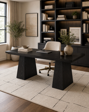 A modern dark wood writing desk with blocky pedestal legs, styled in a home office setting with a cream swivel chair, rug, and a large built-in bookshelf in the background.