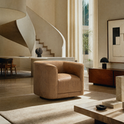 A modern beige armchair in a minimalist, warm-toned interior with a spiral staircase, large windows, and neutral decor.