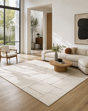 A large off-white area rug with a grid pattern is styled in a bright, modern living room with light wood floors, a cream sectional sofa, and a round wooden coffee table.